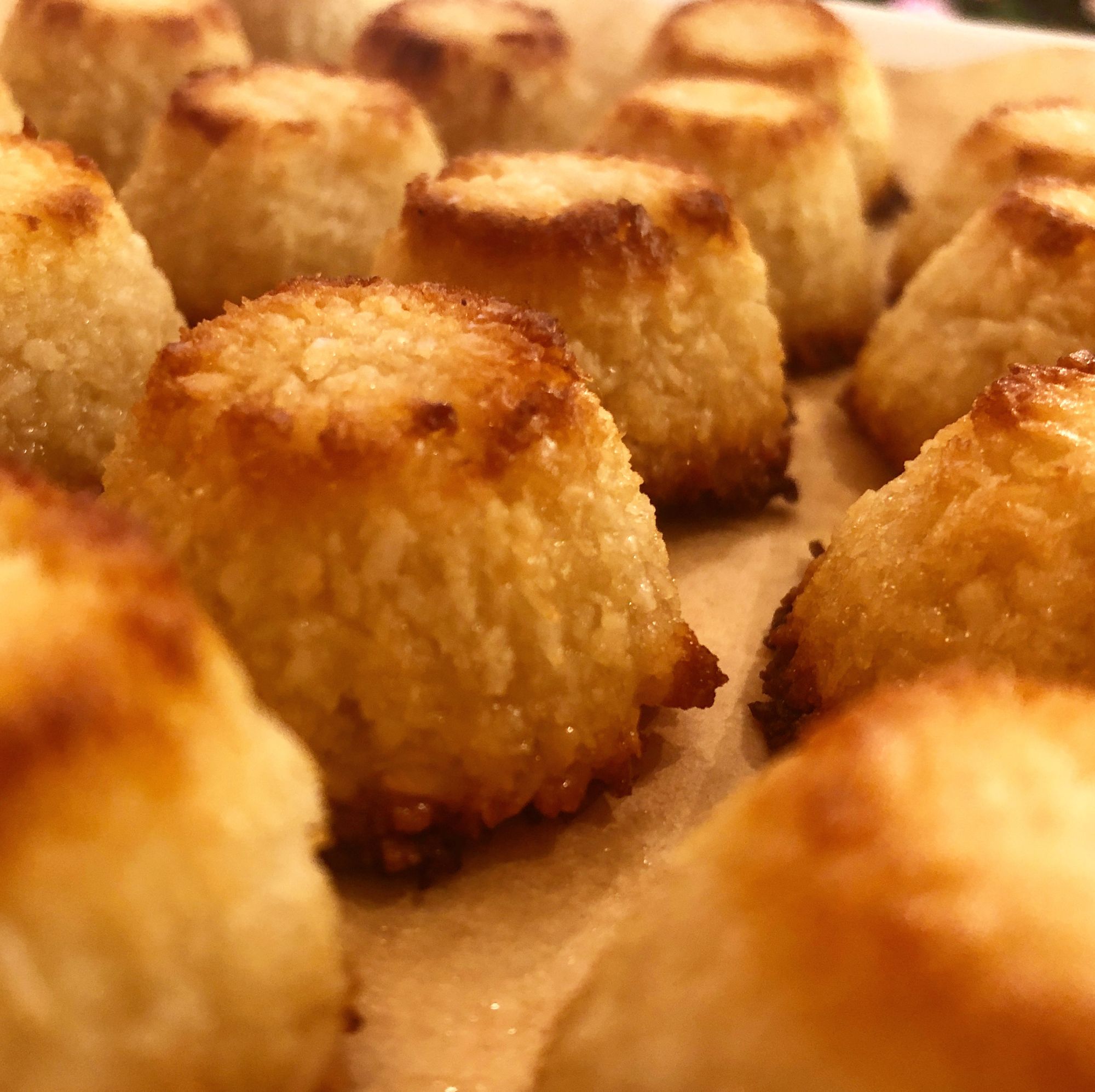 A close-up picture of small lampshade-shaped coconut bites, all golden in colour with a brown ring around the top and bottom edges. They sit on brown parchment paper.